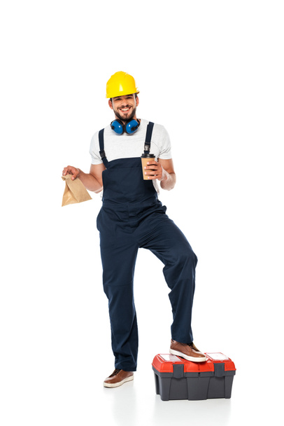 Reparador sonriente en uniforme sosteniendo bolsa de papel y taza desechable cerca de caja de herramientas sobre fondo blanco
 - Foto, Imagen