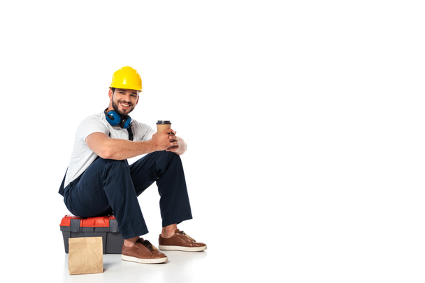 Sorrindo reparador em uniforme segurando café para ir enquanto sentado na caixa de ferramentas perto de saco de papel no fundo branco
 - Foto, Imagem