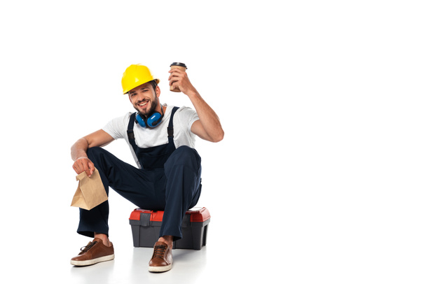 Construtor sorridente segurando café para ir e saco de papel na caixa de ferramentas no fundo branco
 - Foto, Imagem