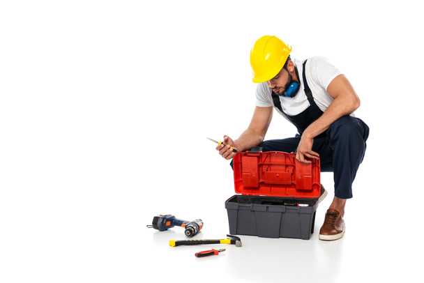 Workman in hardhat and ear defenders holding screwdriver near tools and toolbox on white background - Photo, Image