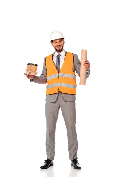 Smiling engineer in suit and safety vest holding paper cups and blueprint tube on white background - Fotó, kép
