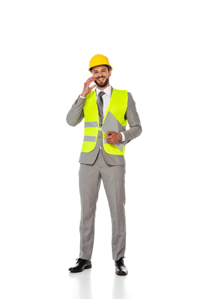 Ingeniero sonriente en traje hablando en el teléfono inteligente y sosteniendo el ordenador portátil sobre fondo blanco
 - Foto, Imagen