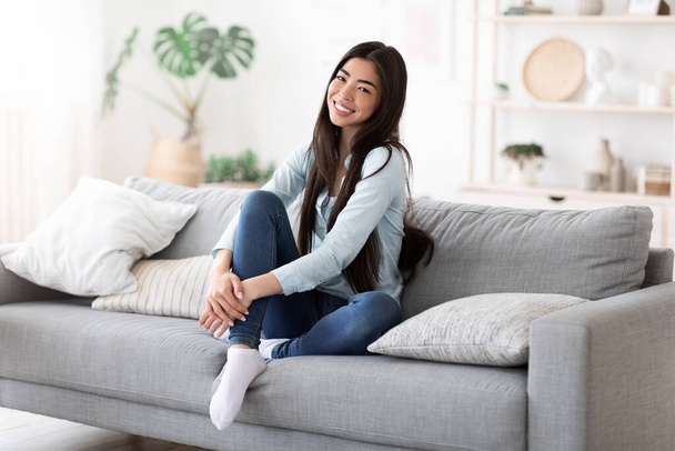 Σπιτική άνεση. Όμορφο χαμογελαστό κορίτσι της Ασίας Posing στον καναπέ στο σαλόνι - Φωτογραφία, εικόνα