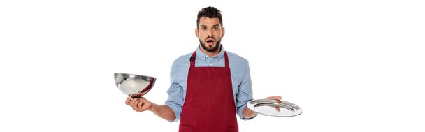 Panoramiczne ujęcie wstrząśniętego kelnera patrzącego w kamerę, trzymającego tacę i pokrywę naczynia na białym tle - Zdjęcie, obraz