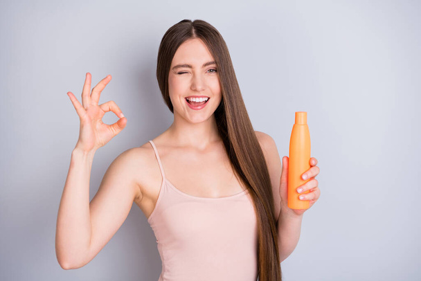Close-up portrait of her she nice-looking attractive pretty cheerful cheery brown-haired girl holding in hand cream bottle showing ok-sign extra length isolated on light gray pastel color background - Foto, afbeelding