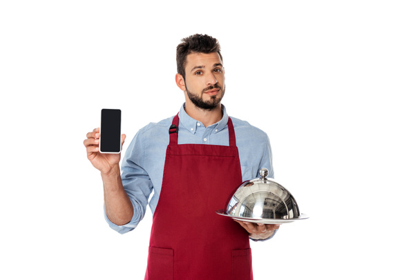 Hübscher Kellner hält Smartphone und Tablett mit Cloche isoliert auf weiß - Foto, Bild