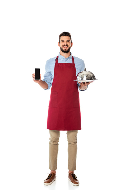 Красивый улыбающийся официант держит смартфон с чистым экраном и поднос с крышкой для тарелки на белом фоне
 - Фото, изображение