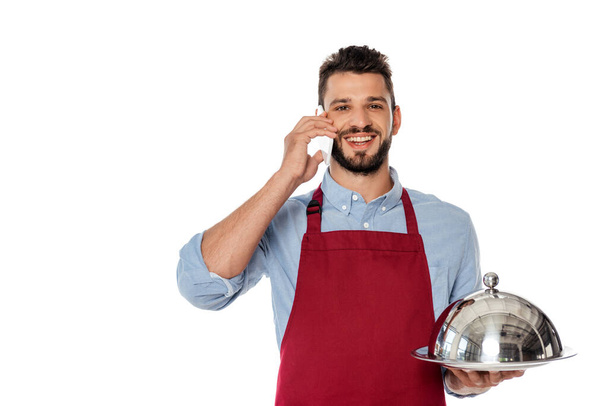 Camarero sonriente hablando en smartphone y sosteniendo bandeja con tapa de plato aislado en blanco
 - Foto, Imagen