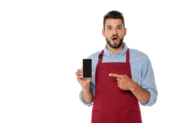 Surprised waiter in apron pointing with finger at smartphone with blank screen isolated on white - Foto, Imagen