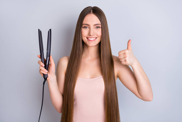 Close-up portrait of her she nice-looking attractive well-groomed cheerful cheery straight-haired girl using straightener showing thumbup advert advice isolated over light gray pastel color background - Фото, зображення
