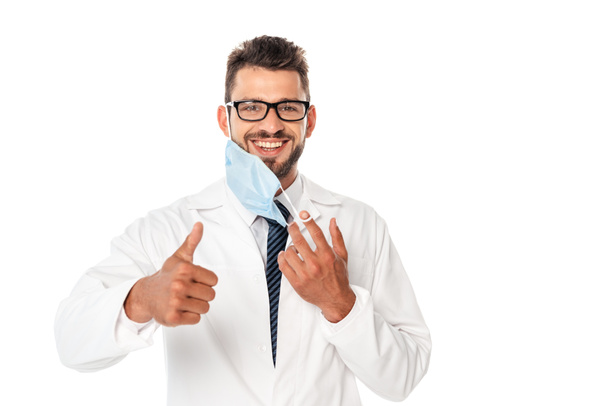 Smiling doctor showing thumb up and holding medical mask isolated on white - Foto, Imagem