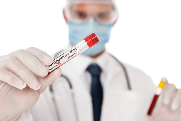 Enfoque selectivo del médico en máscara médica que muestra probeta con muestra de sangre y letras de coronavirus aisladas en blanco
 - Foto, Imagen