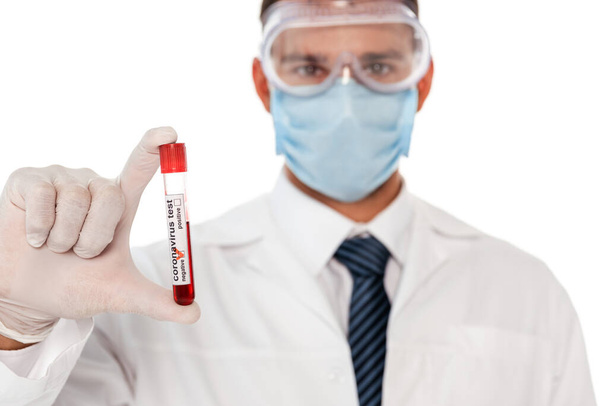 Selective focus of doctor in protective mask and goggles holding test tube with blood sample and coronavirus lettering isolated on white - Фото, изображение