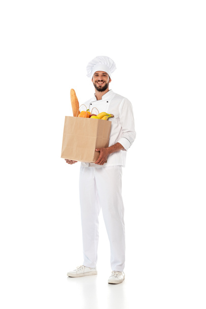 Positive chef holding shopping bag with baguette and fresh fruits on white background - Photo, Image
