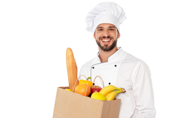 Handsome chef smiling at camera and holding shopping bag with baguette and fresh fruits isolated on white - Foto, immagini