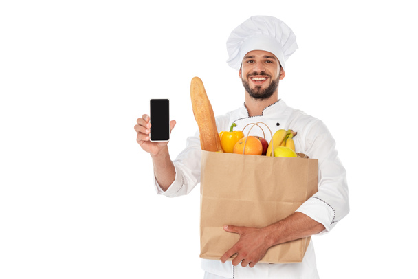 Smiling cook holding shopping bag with food and smartphone isolated on white - Foto, afbeelding