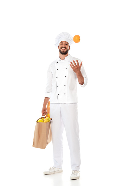 Smiling chef in uniform throwing orange and holding shopping bag with food on white background - Foto, immagini