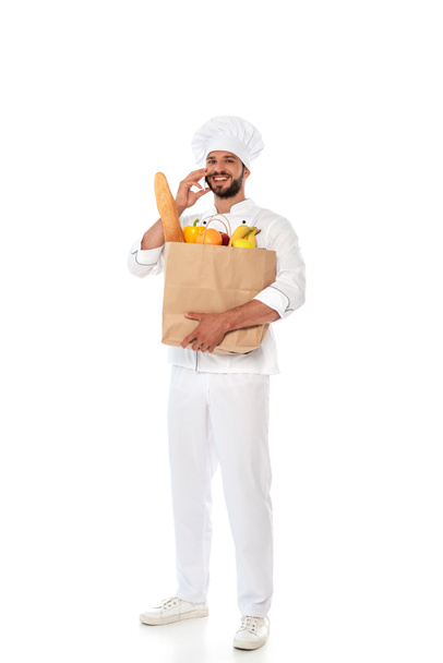 Smiling chef holding shopping bag with food and talking on smartphone on white background - Photo, Image
