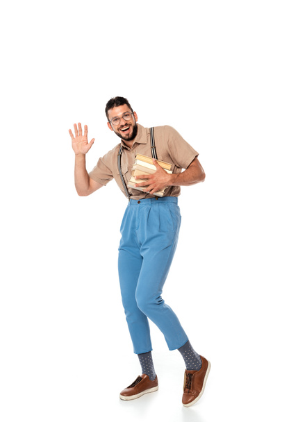 Cheerful nerd in suspenders holding books and waving hand at camera on white background - Φωτογραφία, εικόνα