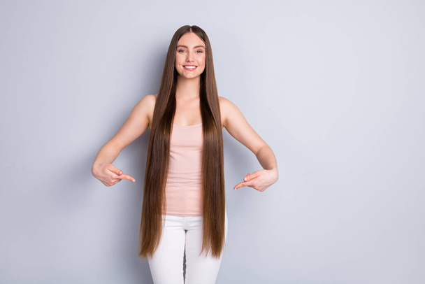 Photo of charming appearance model lady demonstrating ideal neat long hairstyle directing fingers straight hair wear singlet white trousers isolated grey color background - 写真・画像