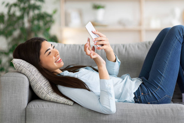 Asiatico ragazza surfing internet su smartphone mentre sdraiato su divano a casa - Foto, immagini
