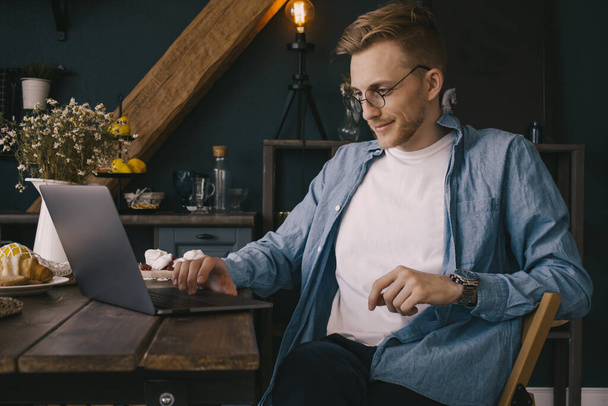 A young guy is sitting at the kitchen table and working on a laptop! A handsome young businessman works from home in a blue shirt - Valokuva, kuva