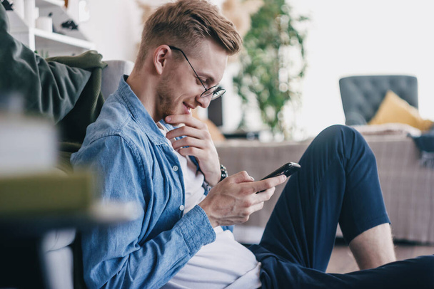 A young man at home with a mobile phone in his hands. Close-up of a young man using the phone at home. A man communicates with friends on a mobile phone. Young business men working from home - 写真・画像