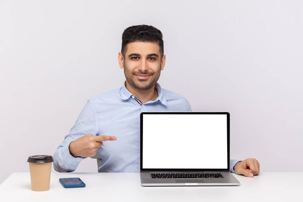 Look at advertisement! Happy elegant man employee sitting in office workplace, pointing laptop blank display, mock up empty place for website promotion. indoor studio shot isolated on white background - Foto, Imagem