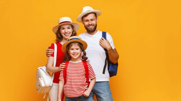 Pozitív szülők és lánya hátizsák mosolyog, és nézi a kamera, miközben a nyári vakáció ellen sárga backgroun - Fotó, kép