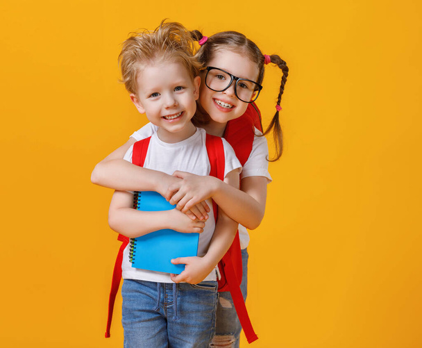 Веселая умная девочка в очках обнимает младшего брата с блокнотом и рюкзаком во время учебы в школе изолированной на желтую спинку
 - Фото, изображение