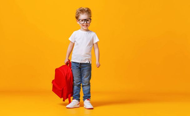Teljes test pozitív kisfiú szemüvegben mosolyog a kamerába és vörös hátizsákot cipel az iskolában a sárga háttér ellen. - Fotó, kép