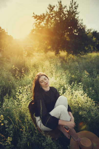 Slow living. Fashionable woman relaxing in summer countryside. Stylish elegant girl sitting on rustic chair in sunset light in summer meadow. Tranquil moment. Creative image. - Photo, Image