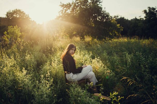 Modische Frau beim Entspannen in der sommerlichen Landschaft. Stilvolle elegante Mädchen sitzen auf rustikalen Stuhl im Sonnenuntergang Licht in Sommerwiese. Atmosphärische ruhigen Moment. Kreatives Image. - Foto, Bild