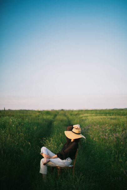 Κομψό κομψό κορίτσι σε ψάθινο καπέλο κάθεται σε ρουστίκ καρέκλα το καλοκαίρι πράσινο λιβάδι στο φως του ήλιου το βράδυ. Μοντέρνα νεαρή γυναίκα χαλαρώνοντας στο πεδίο, γαλήνια στιγμή. - Φωτογραφία, εικόνα
