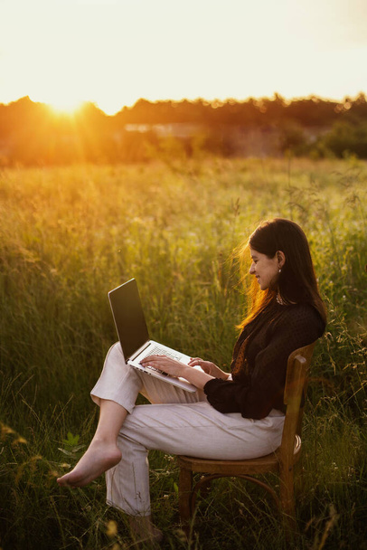 Nová kancelářská koncepce. Módní elegantní dívka pracující na notebooku a sedí na rustikálním křesle ve slunném letním poli při západu slunce. Vzdálená práce se sociálními distancemi a bezpečnostními protokoly - Fotografie, Obrázek