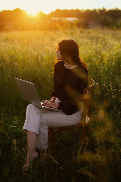 Modisch elegantes Mädchen, das am Laptop arbeitet und auf einem rustikalen Stuhl im sonnigen Sommerfeld bei Sonnenuntergang sitzt. Neues Bürokonzept. Fernarbeit mit sozialer Distanzierung und Sicherheitsprotokollen - Foto, Bild