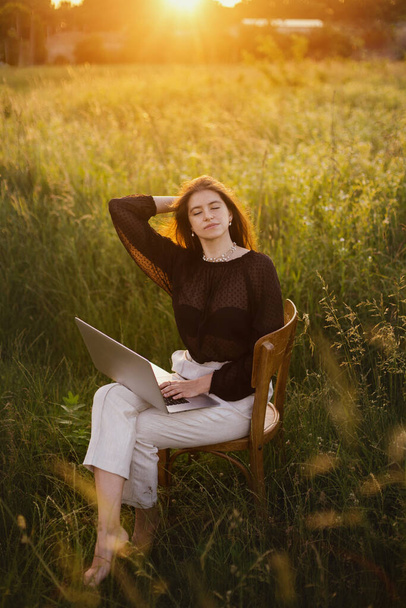 Neues Bürokonzept. Modisch elegantes Mädchen, das am Laptop arbeitet und auf einem rustikalen Stuhl im sonnigen Sommerfeld bei Sonnenuntergang sitzt. Fernarbeit mit sozialer Distanzierung und Sicherheitsprotokollen - Foto, Bild