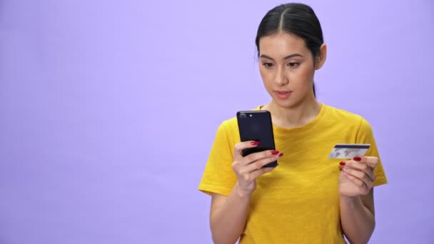 Happy pretty woman in yellow t-shirt paying the bills on smartphone with credit card over violet background - Filmati, video