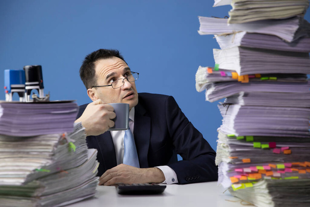 vermoeide kantoormedewerker kijkt bang op hoge stapel documenten - Foto, afbeelding