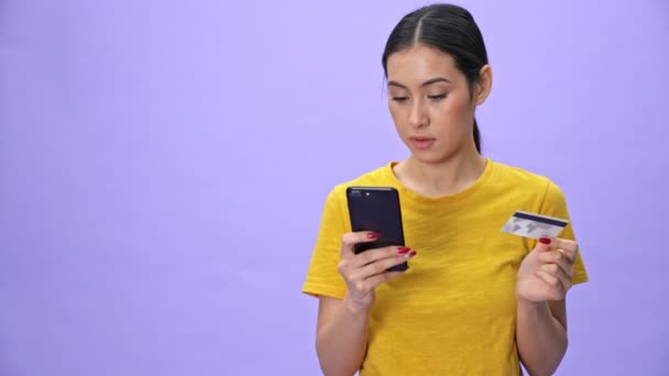Sérieuse jolie femme en t-shirt jaune payant les factures sur smartphone avec carte de crédit sur fond violet - Séquence, vidéo