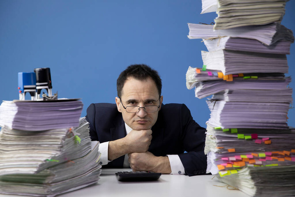 tired office worker puts his head on his fists, amidst high stacks of documents - Photo, Image