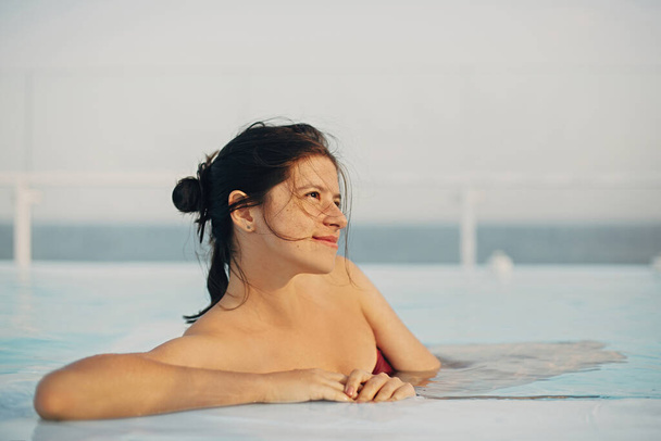 Όμορφη νεαρή γυναίκα χαλαρώνει στην πισίνα, απολαμβάνοντας ζεστό ήλιο και τον άνεμο. Brunette κορίτσι στις καλοκαιρινές διακοπές σε πολυτελές θέρετρο, κολύμπι στην πισίνα στην ταράτσα με θέα στη θάλασσα. Γαληνή στιγμή - Φωτογραφία, εικόνα