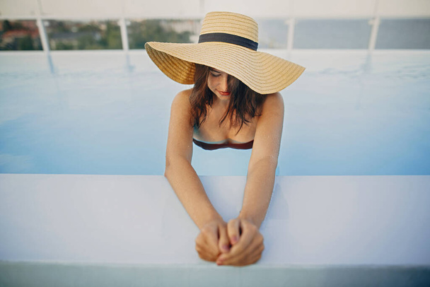 Şapkalı şık genç bir bayan mavi havuzda dinleniyor ve yaz tatilinin tadını çıkarıyor. Yaz tropik tatili. Lüks tatil köyünde güneşli şapkalı bir kız çatıda havuzda yüzüyor. Metin için boşluk - Fotoğraf, Görsel
