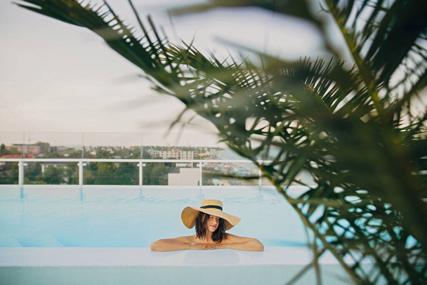 Stylish young woman in hat relaxing in pool under palm leaves and enjoying summer holiday. Summer vacation. Girl in sunhat on vacation in luxury tropical resort, swimming in pool on rooftop - Foto, immagini