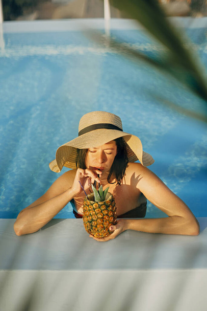 Beautiful young woman in hat drinking delicious cocktail from pineapple and relaxing in pool, summer vacation. Girl enjoying warm sunshine in pool on rooftop in luxury tropical resort - Photo, Image