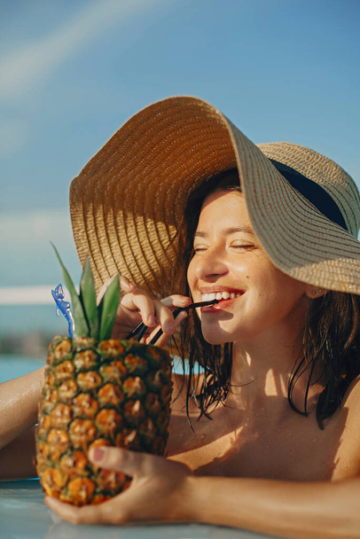 Piękna młoda kobieta delektująca się koktajlem w ananasie, relaksująca się w basenie na słonecznych letnich wakacjach. Portret dziewczyny w kapeluszu z naturalną skórą pijącej koktajl na dachu w luksusowym tropikalnym kurorcie - Zdjęcie, obraz