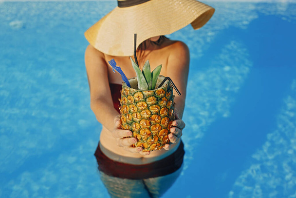 Schöne junge Frau hält Cocktail in Ananas und entspannen im Pool, Sommerferien. Mädchen mit Hut genießt warmen Sonnenschein, schwimmt im Pool auf dem Dach eines luxuriösen tropischen Resorts - Foto, Bild