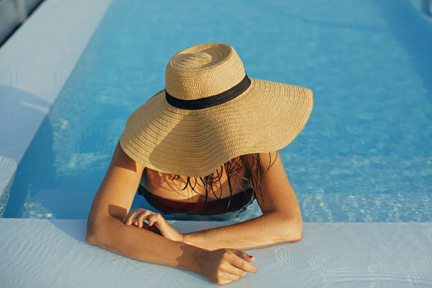 青いプールでリラックスし、夏休みを楽しむ帽子のスタイリッシュな若い女性。夏の熱帯休暇。豪華なリゾートで休暇中のサンハットの女の子は、屋上のプールで泳いでいます。テキストのスペース - 写真・画像