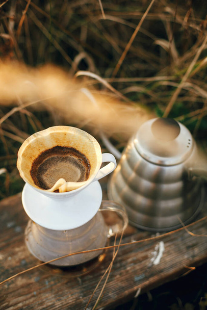 屋外での代替コーヒー醸造。大気素朴な瞬間。夕日の田園地帯を背景に、フィルターやコーヒー泡が付いたガラス製のフラスコドリッパーに湯を入れる. - 写真・画像