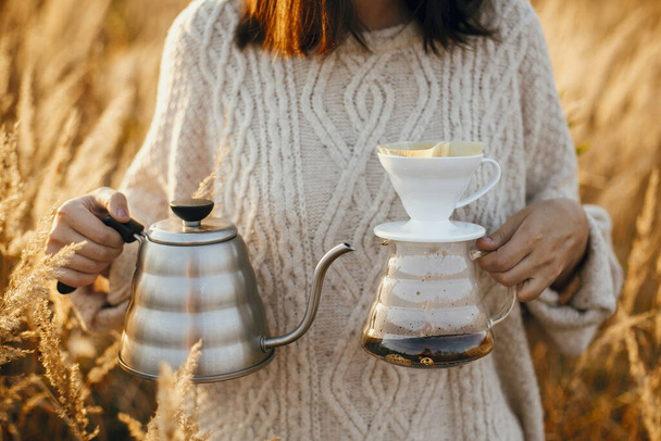 Alternatif kahve açık havada güneşli bir ışıkta demleniyor. Günbatımında kırsal otların arka planında filtreli sıcak su ısıtıcısı ve cam damlası tutan hippi gezgin. Atmosferik kırsal an - Fotoğraf, Görsel